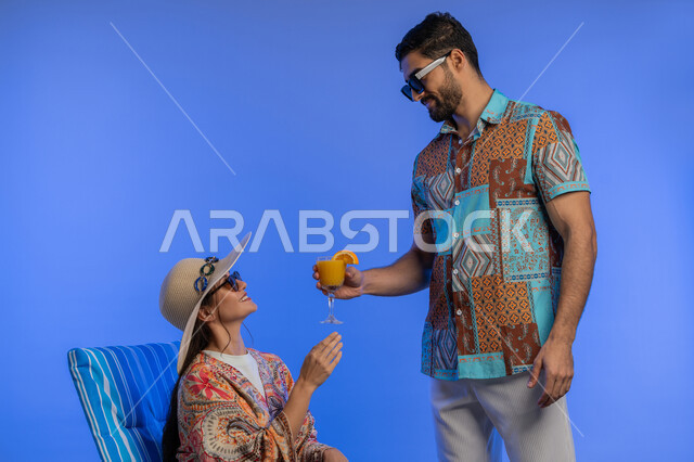 Tourism, recreation and spending a pleasant summer time, a portrait of a smiling young Arab Gulf Emirati woman wearing casual clothes, sunglasses and a straw hat sitting on a comfortable chair enjoying a tourist trip, a Saudi man offering his wife refreshing natural juice, blue background