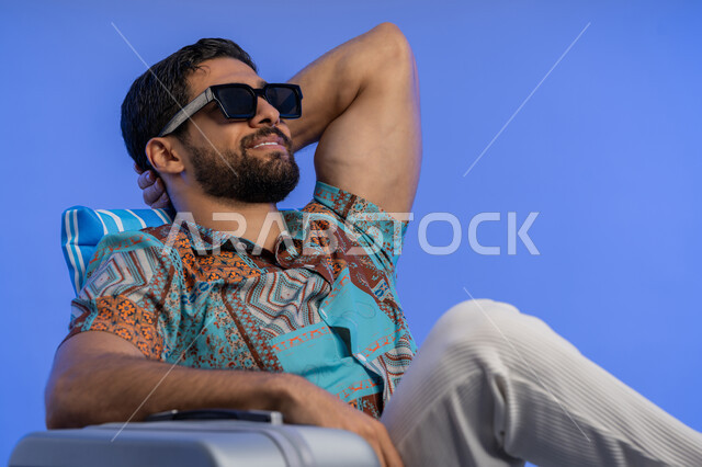الرحلات السياحية في المنتجعات والشواطئ, بورتريه مقرب لشاب عربي خليجي اماراتي مبتسم يرتدي الزي الكاجوال والنظارة الشمسية يجلس على كرسي مريح, رجل سعودي يقوم بالنظر إلى شيء ما بإيماءات الاسترخاء والراحة, الاستمتاع بالإجازة والمناظر الطبيعيه, خلفية زرقاء