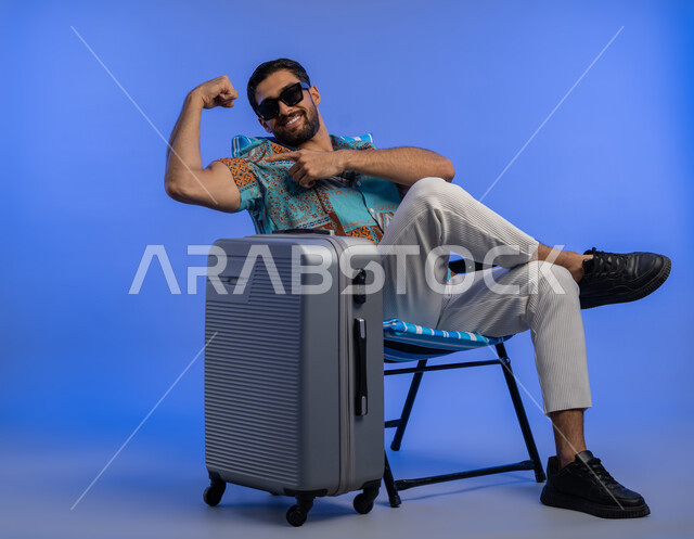 استعراض القوة البدنية, بورتريه لشاب عربي خليجي اماراتي مبتسم يرتدي الزي الكاجوال والنظارة الشمسية يرفع يده بكفة مقبوضة, رجل سعودي يعرض العضلات بإيماءات الثقة والسرور, أهمية السفر وقضاء أوقات ممتعة في الأماكن السياحية, خلفية زرقاء