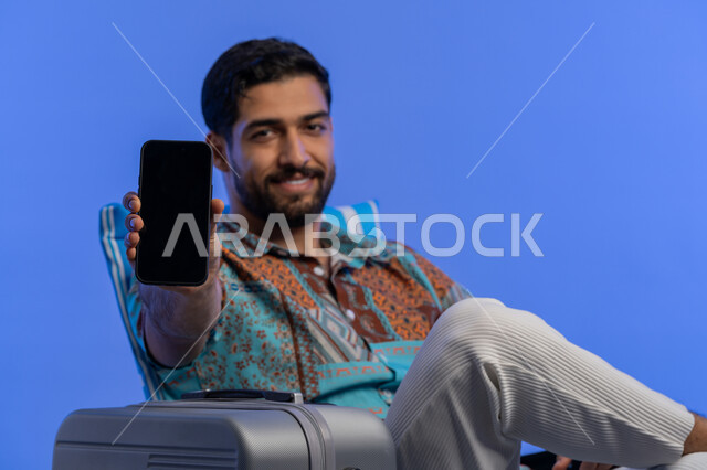 Displaying tourism and travel services and applications, an enjoyable youth trip and an entertaining summer vacation, a portrait of a smiling Saudi Arabian Gulf young man wearing casual clothes sitting next to a bag displaying a mobile phone with a blank black screen, using modern and advanced devices and technologies, blue background