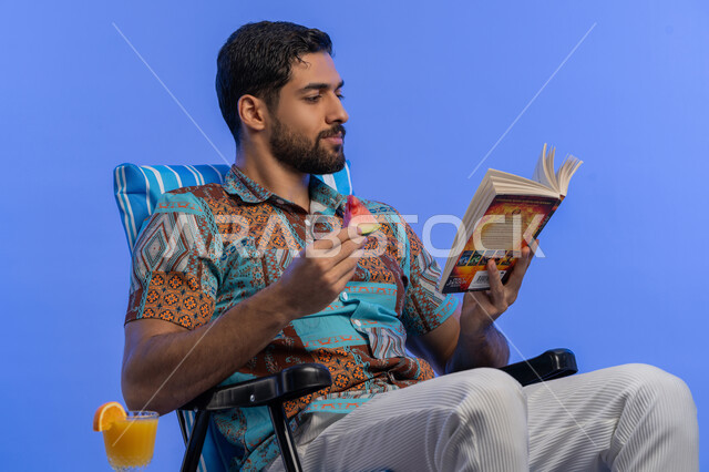 تناول الفواكه الصيفية المنعشة, ممارسة هواية مفيدة في أوقات الفراغ, بورتريه مقرب لشاب عربي خليجي اماراتي مبتسم يرتدي الزي الكاجوال يقوم بقراءة كتاب ما, رحلة شابية ممتعه, قضاء وقت العطلة الصيفية بالسياحة, رجل سعودي يجلس على كرسي مريح بتعابير السرور, خلفية زرقاء