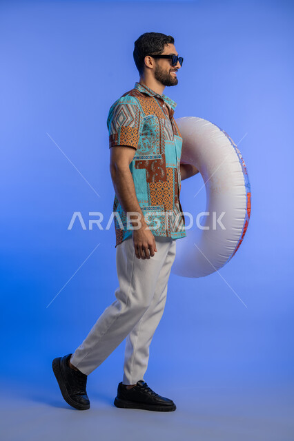 ايماءات الاستعداد للذهاب والمشي, بورتريه من الجانب لشاب عربي خليجي اماراتي مبتسم يرتدي الزي الكاجوال والنظارة الشمسية يمسك بيده عوامة السباحة, رجل سعودي بتعابير السرور والسعادة, قضاء وقت ممتع في فصل الصيف, الاستمتاع برحلات العطلة, الجسم بالطول الكامل, خلفية زرقاء
