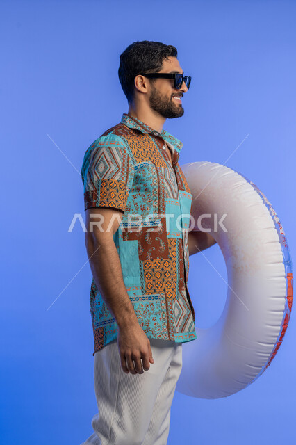 ايماءات الاستعداد للذهاب والمشي, بورتريه من الجانب لشاب عربي خليجي اماراتي مبتسم يرتدي الزي الكاجوال والنظارة الشمسية يمسك بيده عوامة السباحة, رجل سعودي بتعابير السرور والسعادة, قضاء وقت ممتع في فصل الصيف, الاستمتاع برحلات العطلة, خلفية زرقاء