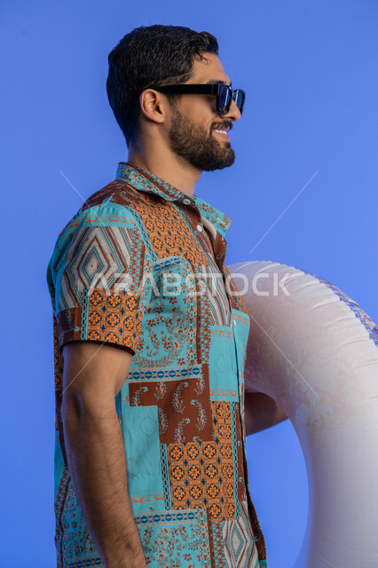ايماءات الاستعداد للذهاب والمشي, بورتريه مقرب من الجانب لشاب عربي خليجي اماراتي مبتسم يرتدي الزي الكاجوال والنظارة الشمسية يمسك بيده عوامة السباحة, رجل سعودي بتعابير السرور والسعادة, قضاء وقت ممتع في فصل الصيف, الاستمتاع برحلات العطلة, خلفية زرقاء
