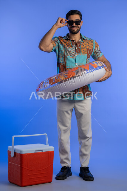 قضاء وقت ممتع في فصل الصيف, بورتريه لشاب عربي خليجي اماراتي مبتسم يرتدي الزي الكاجوال والنظارة الشمسية ينظر إلى الكاميرا بتعابير السرور, رجل سعودي يرتدي العوامة يستعد للسباحة في الشاطئ, حافظة البرودة للعصائر, الاستمتاع بالعطلة الصيفية, الجسم بالطول الكامل, خلفية زرقاء