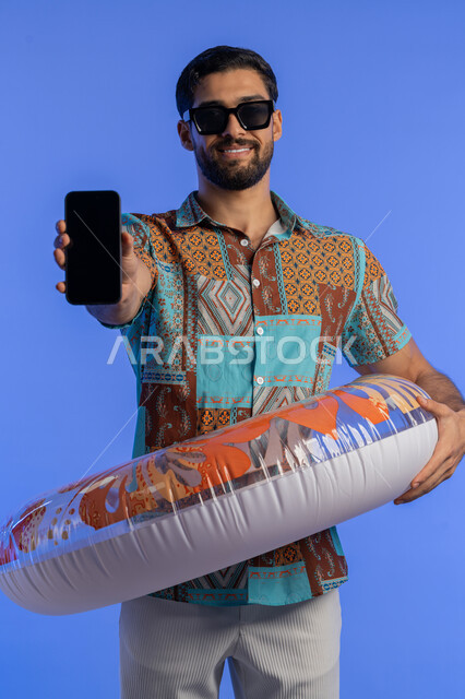 عرض تطبيقات خدمات الأنشطة السياحية, بورتريه لشاب عربي خليجي اماراتي مبتسم يرتدي الزي الكاجوال وعوامة السباحة يحمل الهاتف المحمول بشاشة سوداء فارغة, قضاء وقت ممتع بالعطلة الصيفية, رجل سعودي بتعابير التسليه والترفيه في إحدى المنتجعات, خلفية زرقاء