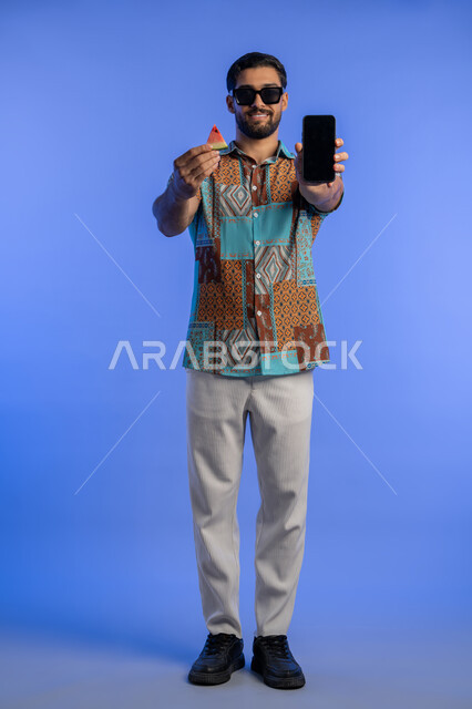 تناول الفواكه الصيفية المنعشة, رجل سعودي يستخدم الهاتف المحمول, بورتريه لرجل عربي خليجي اماراتي مبتسم يرتدي الزي الكاجوال والنظارة الشمسية يعرض الجوال بشاشة سوداء فارغة, الاستمتاع بالإجازة السياحية بإيماءات السرور والبشاشة, الجسم بالطول الكامل, خلفية زرقاء