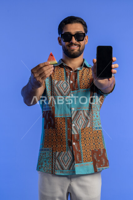 تناول الفواكه الصيفية المنعشة, رجل سعودي يستخدم برامج وتطبيقات الهاتف المحمول, بورتريه لرجل عربي خليجي اماراتي مبتسم يرتدي الزي الكاجوال والنظارة الشمسية يعرض الجوال بشاشة سوداء فارغة, الاستمتاع بالإجازة السياحية بإيماءات السرور والبشاشة, خلفية زرقاء