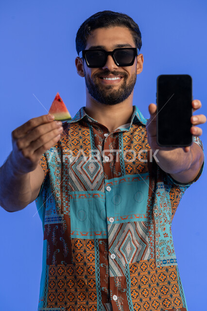 تناول الفواكه الصيفية المنعشة, رجل سعودي يستخدم برامج وتطبيقات الهاتف المحمول, بورتريه مقرب لرجل عربي خليجي اماراتي مبتسم يرتدي الزي الكاجوال والنظارة الشمسية يعرض الجوال بشاشة سوداء فارغة, الاستمتاع بالإجازة السياحية بإيماءات السرور والبشاشة, خلفية زرقاء