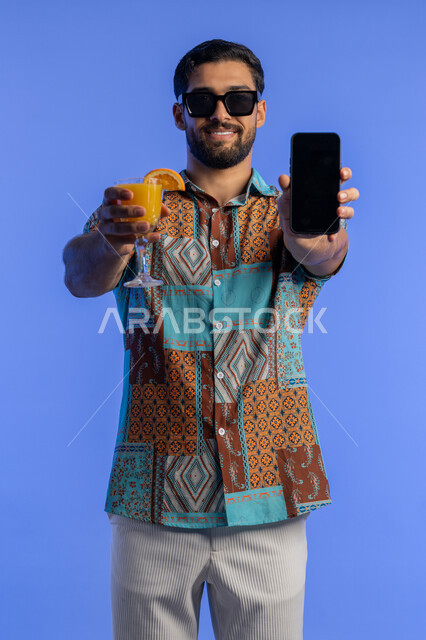 عرض خدمات وتطبيقات السياحة والسفر, رجل سعودي يستمتع بشرب العصير الطبيعي البارد, رحلة شبابية ممتعه وقضاء وقت مسلي في العطلة الصيفية, بورتريه لشاب عربي خليجي اماراتي مبتسم يرتدي الزي الكاجوال والنظارة الشمسية يعرض هاتف محمول بشاشة سوداء فارغة, خلفية زرقاء