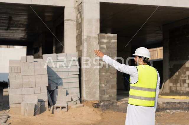 الاشارة الى شيء ما، مهنة هندسية شبابية، مهندس عربي خليجي اماراتي يرتدي الكندورة والسترة وخوذة الحماية يعمل في القطاع الهندسي في الامارات العربية المتحدة، الاشرف على عمليات البناء، مفهوم ادارة المشاريع الهندسية والانشاءات، دراسة المخطط والمشروع على ارض الواقع