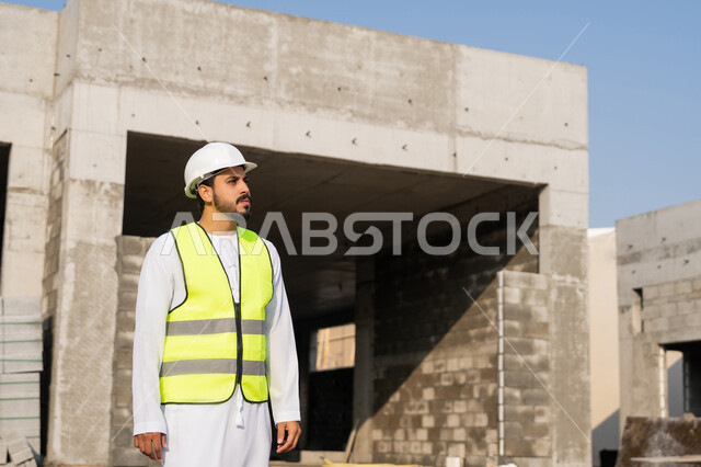 The concept of engineering and construction project management, studying the plan and the project on the ground, a youth engineering profession and job, an Arab Gulf Emirati engineer wearing a kandura, jacket and protective helmet, working in the engineering sector in the United Arab Emirates, supervising construction operations.