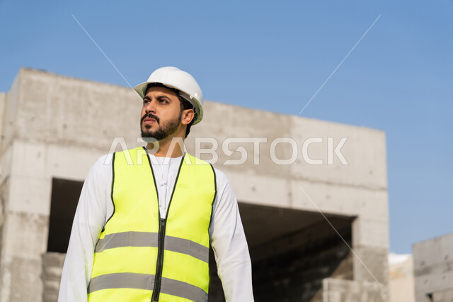 مفهوم ادارة المشاريع الهندسية والانشاءات، دراسة المخطط والمشروع على ارض الواقع، مهنة ووظيفة هندسية شبابية، مهندس عربي خليجي اماراتي يرتدي الكندورة والسترة وخوذة الحماية يعمل في القطاع الهندسي في الامارات العربية المتحدة، الاشرف على عمليات البناء