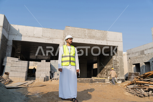 The concept of engineering and construction project management, studying the plan and the project on the ground, a youth engineering profession and job, an Arab Gulf Emirati engineer wearing a kandura, jacket and protective helmet, working in the engineering sector in the United Arab Emirates, supervising construction operations.