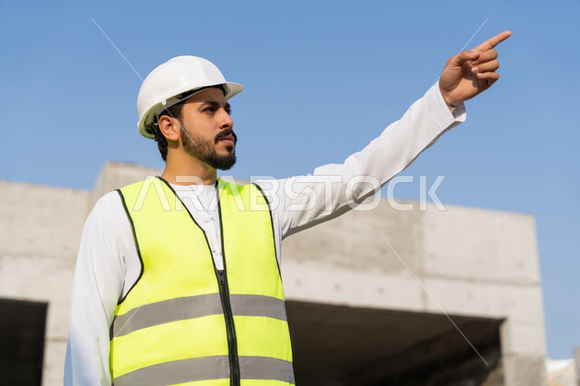 الاشارة الى شيء ما، مهنة هندسية شبابية، مهندس عربي خليجي اماراتي يرتدي الكندورة والسترة وخوذة الحماية يعمل في القطاع الهندسي في الامارات العربية المتحدة، الاشرف على عمليات البناء، مفهوم ادارة المشاريع الهندسية والانشاءات، دراسة المخطط والمشروع على ارض الواقع
