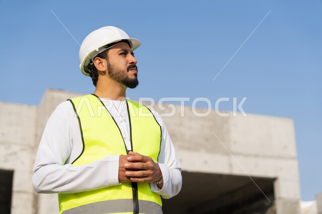 مفهوم ادارة المشاريع الهندسية والانشاءات، دراسة المخطط والمشروع على ارض الواقع، مهنة ووظيفة هندسية شبابية، مهندس عربي خليجي اماراتي يرتدي الكندورة والسترة وخوذة الحماية يعمل في القطاع الهندسي في الامارات العربية المتحدة، الاشرف على عمليات البناء