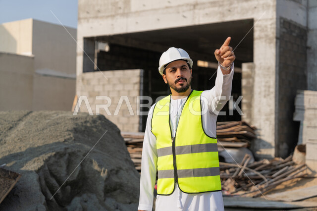 الاشارة الى شيء ما، مهنة هندسية شبابية، مهندس عربي خليجي اماراتي يرتدي الكندورة والسترة وخوذة الحماية يعمل في القطاع الهندسي في الامارات العربية المتحدة، الاشرف على عمليات البناء، مفهوم ادارة المشاريع الهندسية والانشاءات، دراسة المخطط والمشروع على ارض الواقع