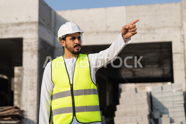 الاشارة الى شيء ما، مهنة هندسية شبابية، مهندس عربي خليجي اماراتي يرتدي الكندورة والسترة وخوذة الحماية يعمل في القطاع الهندسي في الامارات العربية المتحدة، الاشرف على عمليات البناء، مفهوم ادارة المشاريع الهندسية والانشاءات، دراسة المخطط والمشروع على ارض الواقع
