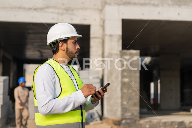 استخدام جهاز تقني تكنولوجي، مهنة هندسية شبابية، مهندس عربي خليجي اماراتي يرتدي الكندورة والسترة والخوذة يمسك الهاتف المحمول، العمل في القطاع الهندسي في الامارات، الاشرف على عمليات البناء، ادارة المشاريع الهندسية والانشاءات، دراسة المخطط والمشروع على ارض الواقع