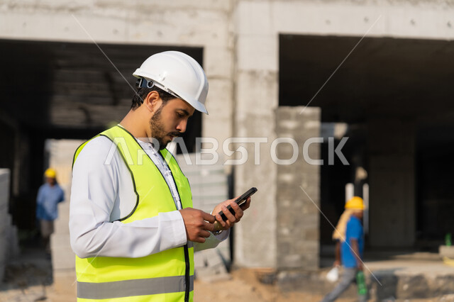 استخدام جهاز تقني تكنولوجي، مهنة هندسية شبابية، مهندس عربي خليجي اماراتي يرتدي الكندورة والسترة والخوذة يمسك الهاتف المحمول، العمل في القطاع الهندسي في الامارات، الاشرف على عمليات البناء، ادارة المشاريع الهندسية والانشاءات، دراسة المخطط والمشروع على ارض الواقع