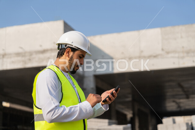 استخدام جهاز تقني تكنولوجي، مهنة هندسية شبابية، مهندس عربي خليجي اماراتي يرتدي الكندورة والسترة والخوذة يمسك الهاتف المحمول، العمل في القطاع الهندسي في الامارات، الاشرف على عمليات البناء، ادارة المشاريع الهندسية والانشاءات، دراسة المخطط والمشروع على ارض الواقع