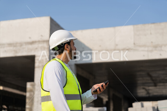استخدام جهاز تقني تكنولوجي، مهنة هندسية شبابية، مهندس عربي خليجي اماراتي يرتدي الكندورة والسترة والخوذة يمسك الهاتف المحمول، العمل في القطاع الهندسي في الامارات، الاشرف على عمليات البناء، ادارة المشاريع الهندسية والانشاءات، دراسة المخطط والمشروع على ارض الواقع