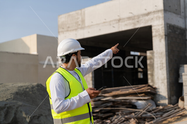 Using a technological device, a youth engineering profession, an Arab Gulf Emirati engineer wearing a kandura, jacket and helmet holding a mobile phone, working in the engineering sector in the Emirates, supervising construction operations, managing engineering and construction projects, studying the plan and the project on the ground