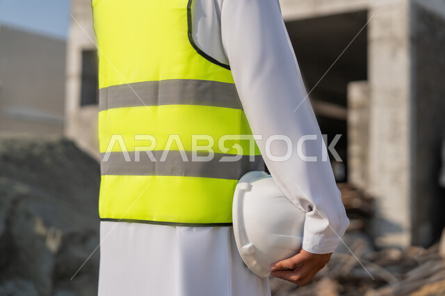 متابعة أعمال البناء وصب الخرسانات ووضع الأساسات الأولية، مهنة ووظيفة شبابية هندسية، صورة مقربة من الخلف لمهندس عربي خليجي اماراتي يرتدي السترة يمسك الخوذة يقوم بالاشراف على مبنى قيد الإنشاء والتشييد في موقع البناء، مفهوم الهندسة الانشائية والمعمارية