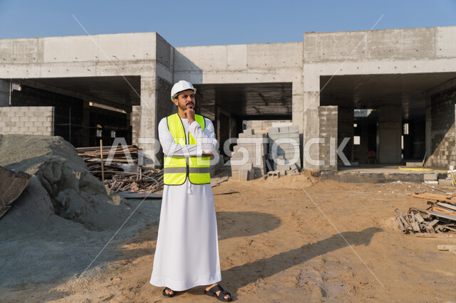 Standing with expressions of thought and absent-mindedness, observing construction work, pouring concrete and laying the initial foundations, a youth engineering profession and job, an Arab Gulf Emirati engineer wearing a kandura, jacket and helmet supervising a building under construction and erection, the concept of structural and architectural engineering