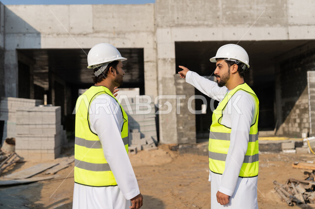 مفهوم ادارة المشاريع الهندسية والانشاءات، دراسة المخطط والمشروع على ارض الواقع، مهنة ووظيفة هندسية شبابية، مهندسان عربيان خليجيان اماراتيان يرتديان الكندورة والسترة وخوذة الحماية يعملان في القطاع الهندسي في الامارات، مراقبة ومتابعة عمليات البناء