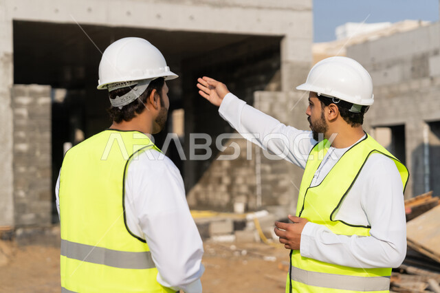 مراقبة ومتابعة عمليات البناء، مفهوم ادارة المشاريع الهندسية والانشاءات، دراسة المخطط والمشروع على ارض الواقع، مهنة ووظيفة هندسية شبابية، مهندسان عربيان خليجيان اماراتيان يرتديان الكندورة والسترة وخوذة الحماية يعملان في القطاع الهندسي في الامارات