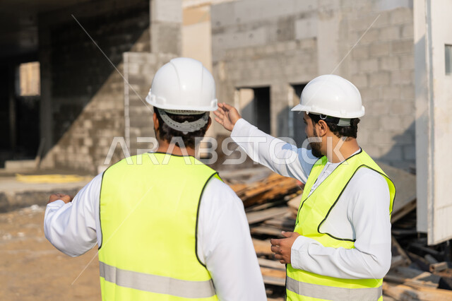 دراسة المخطط والمشروع على ارض الواقع، مهنة ووظيفة هندسية شبابية، مهندسان عربيان خليجيان اماراتيان يرتديان الكندورة والسترة وخوذة الحماية يعملان في القطاع الهندسي في الامارات، مراقبة ومتابعة عمليات البناء، مفهوم ادارة المشاريع الهندسية والانشاءات