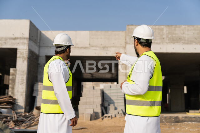 تبادل المعلومات والخبرة، دراسة المخطط والمشروع على ارض الواقع، وظيفة هندسية شبابية، مهندسان عربيان خليجيان اماراتيان يرتديان الكندورة والسترة وخوذة الحماية يعملان في القطاع الهندسي في الامارات، مراقبة ومتابعة عمليات البناء، مفهوم ادارة المشاريع الهندسية والانشاءات