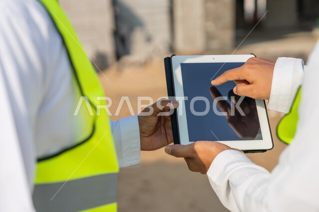 Using a technical tablet in the engineering field, studying the plan on the ground, a youth engineering job, two Arab Gulf Emirati engineers wearing kanduras, jackets and protective helmets, monitoring and following up on construction operations, managing engineering and construction projects using technology.