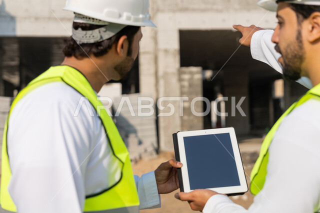 Using a technical tablet in the engineering field, studying the plan on the ground, a youth engineering job, two Arab Gulf Emirati engineers wearing kanduras, jackets and protective helmets, monitoring and following up on construction operations, managing engineering and construction projects using technology.