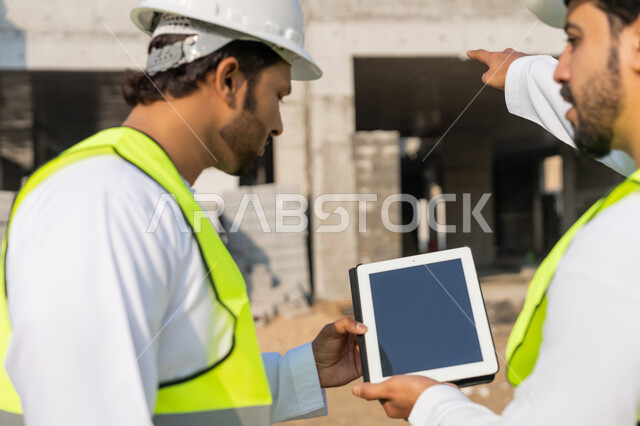 استخدام جهاز التابلت التقني في المجال الهندسي، دراسة المخطط على ارض الواقع، وظيفة هندسية شبابية، مهندسان عربيان خليجيان اماراتيان يرتديان الكندورة والسترة وخوذة الحماية يقومان لمراقبة ومتابعة عمليات البناء، ادارة المشاريع الهندسية والانشاءات باستخدام التكنولوجيا