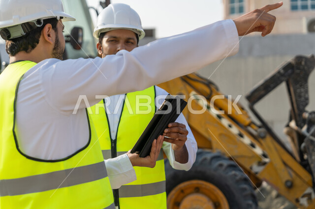 Using a technical tablet in the engineering field, studying the plan on the ground, a youth engineering job, two Arab Gulf Emirati engineers wearing kanduras, jackets and protective helmets, monitoring and following up on construction operations, managing engineering and construction projects using technology.