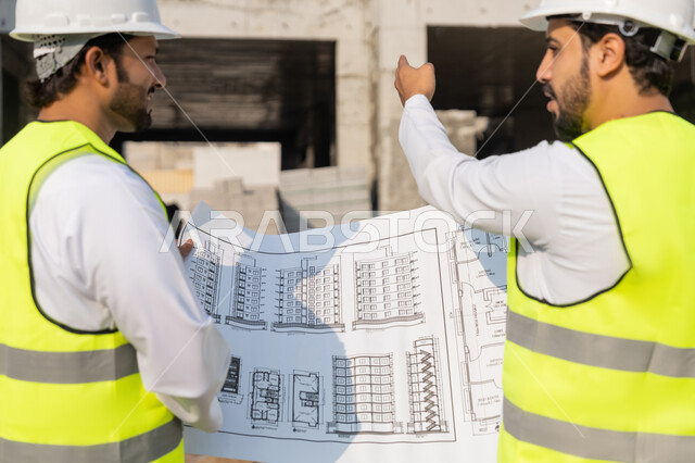 تبادل المعلومات والخبرة، دراسة المخطط والمشروع على ارض الواقع، وظيفة هندسية شبابية، مهندسان عربيان خليجيان اماراتيان يرتديان الكندورة والسترة وخوذة الحماية يعملان في القطاع الهندسي في الامارات، مراقبة ومتابعة عمليات البناء، مفهوم ادارة المشاريع الهندسية والانشاءات