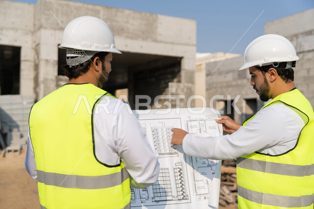 مراقبة ومتابعة عمليات البناء، مفهوم ادارة المشاريع الهندسية والانشاءات، دراسة المخطط والمشروع على ارض الواقع، مهنة ووظيفة هندسية شبابية، مهندسان عربيان خليجيان اماراتيان يرتديان الكندورة والسترة وخوذة الحماية يعملان في القطاع الهندسي في الامارات