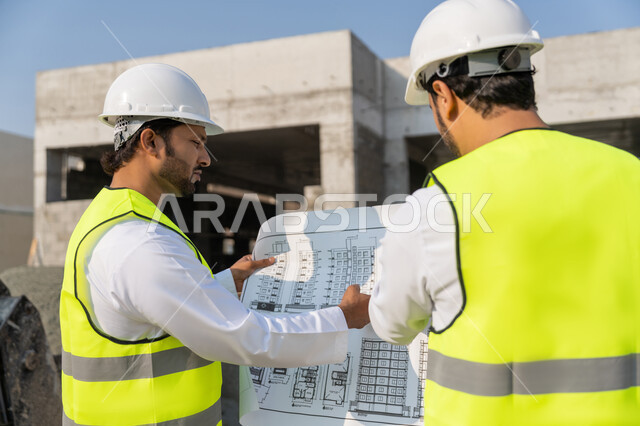 مفهوم ادارة المشاريع الهندسية والانشاءات، دراسة المخطط والمشروع على ارض الواقع، مهنة ووظيفة هندسية شبابية، مهندسان عربيان خليجيان اماراتيان يرتديان الكندورة والسترة وخوذة الحماية يعملان في القطاع الهندسي في الامارات، مراقبة ومتابعة عمليات البناء