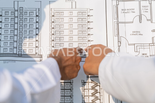 دراسة المخطط والمشروع على ارض الواقع، وظيفة هندسية شبابية، صورة مقربة من الخلف لمهندسان عربيان خليجيان اماراتيان يرتديان الكندورة والسترة يشيران الى المخطط، العمل في القطاع الهندسي في الامارات، مراقبة ومتابعة عمليات البناء، مفهوم ادارة المشاريع الهندسية والانشاءات