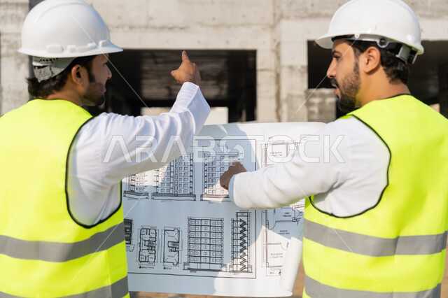 Exchange of information and experience, study of the plan and project on the ground, a youth engineering job, two Arab Gulf Emirati engineers wearing kandura, jacket and protective helmet working in the engineering sector in the Emirates, monitoring and following up on construction operations, the concept of engineering and construction project management.