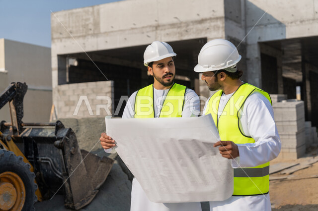 مراقبة ومتابعة عمليات البناء، مفهوم ادارة المشاريع الهندسية والانشاءات، دراسة المخطط والمشروع على ارض الواقع، مهنة ووظيفة هندسية شبابية، مهندسان عربيان خليجيان اماراتيان يرتديان الكندورة والسترة وخوذة الحماية يعملان في القطاع الهندسي في الامارات