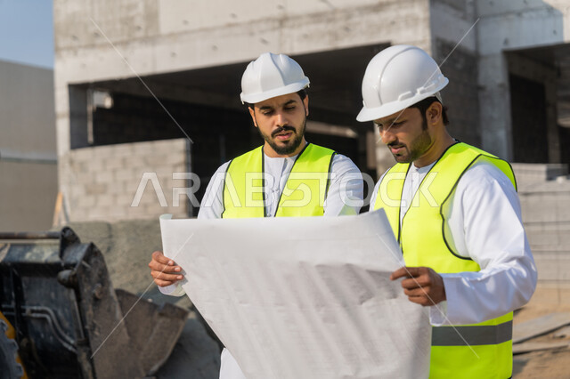 The concept of engineering and construction project management, studying the plan and the project on the ground, a youth engineering profession and job, two Arab Gulf Emirati engineers wearing kanduras, jackets and protective helmets working in the engineering sector in the Emirates, monitoring and following up on construction operations.