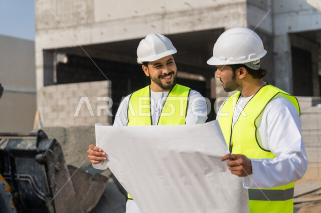 دراسة المخطط والمشروع على ارض الواقع، مهنة ووظيفة هندسية شبابية، مهندسان عربيان خليجيان اماراتيان يرتديان الكندورة والسترة وخوذة الحماية يعملان في القطاع الهندسي في الامارات، مراقبة ومتابعة عمليات البناء، مفهوم ادارة المشاريع الهندسية والانشاءات