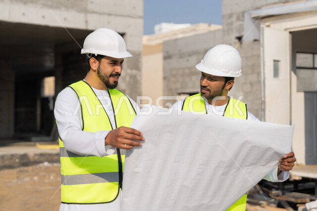 مراقبة ومتابعة عمليات البناء، مفهوم ادارة المشاريع الهندسية والانشاءات، دراسة المخطط والمشروع على ارض الواقع، مهنة ووظيفة هندسية شبابية، مهندسان عربيان خليجيان اماراتيان يرتديان الكندورة والسترة وخوذة الحماية يعملان في القطاع الهندسي في الامارات