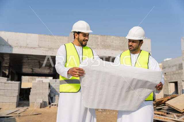 مفهوم ادارة المشاريع الهندسية والانشاءات، دراسة المخطط والمشروع على ارض الواقع، مهنة ووظيفة هندسية شبابية، مهندسان عربيان خليجيان اماراتيان يرتديان الكندورة والسترة وخوذة الحماية يعملان في القطاع الهندسي في الامارات، مراقبة ومتابعة عمليات البناء