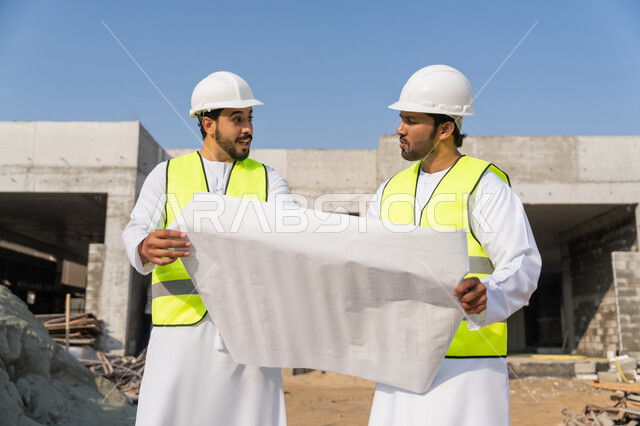 مراقبة ومتابعة عمليات البناء، مفهوم ادارة المشاريع الهندسية والانشاءات، دراسة المخطط والمشروع على ارض الواقع، مهنة ووظيفة هندسية شبابية، مهندسان عربيان خليجيان اماراتيان يرتديان الكندورة والسترة وخوذة الحماية يعملان في القطاع الهندسي في الامارات