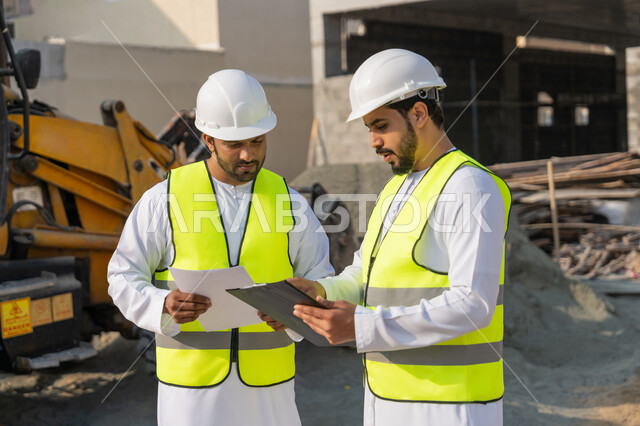 دراسة المخطط والمشروع على ارض الواقع، مهنة ووظيفة هندسية شبابية، مهندسان عربيان خليجيان اماراتيان يرتديان الكندورة والسترة وخوذة الحماية يعملان في القطاع الهندسي في الامارات، مراقبة ومتابعة عمليات البناء، مفهوم ادارة المشاريع الهندسية والانشاءات