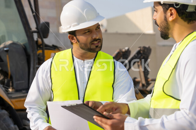 Exchange of information and experience, study of the plan and project on the ground, a youth engineering job, two Arab Gulf Emirati engineers wearing kandura, jacket and protective helmet working in the engineering sector in the Emirates, monitoring and following up on construction operations, the concept of engineering and construction project management.